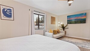 A bedroom with a large bed, a sofa with cushions, a window with curtains, a painting on the wall, and a ceiling fan. - Badger Den - Walk to Village, Walk to Shops, Modern, Fenced Yard (Big Bear Lake)