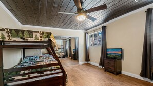 A bedroom with a bunk bed, wooden flooring, a ceiling fan, a TV, and a window with curtains.