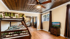 A room with a bunk bed, wooden flooring, a ceiling fan, a television, and a window with curtains. - Jaybird Lodge - Walk to Lake & Village, Fireplace, AC (Big Bear Lake)