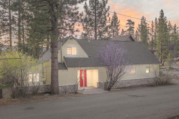 A two-story house with a red door, surrounded by trees and a stone wall.