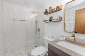A modern bathroom with a herringbone tile backsplash, a glass-enclosed shower, a white toilet, and a vanity with a gold faucet.