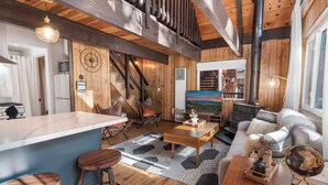 A spacious living area with a wooden ceiling, a large window, a sofa, a coffee table, and a kitchen area with a marble countertop and stools.