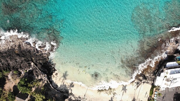 Eine Luftaufnahme eines Strandes mit klarem türkisblauem Wasser, felsiger Küstenlinie und einigen Gebäuden mit Außenbereichen.