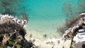 Eine Vogelperspektive zeigt einen Strand mit klarem türkisblauem Wasser, einer felsigen Küstenlinie und einem Resortbereich mit Gebäuden und Palmen.