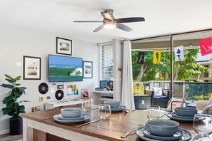 A dining area with a wooden table set for four, a television mounted on the wall, and a ceiling fan. - Modern Kona Retreat • Walk to Ocean & Town (Kailua-Kona)