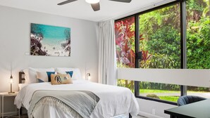 Ein Schlafzimmer mit Bett, Deckenventilator, Nachttisch, Tisch und einem großen Fenster mit Blick auf Grünflächen.