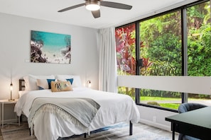 A bedroom with a bed, a ceiling fan, a nightstand, a table, and a large window with a view of greenery. - Modern Kona Retreat • Walk to Ocean & Town (Kailua-Kona)