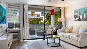 Ein modernes Wohnzimmer mit einem weißen Sofa, einem Glastisch und einem Deckenventilator. Ein Schiebetür aus Glas führt auf einen Balkon mit Gartenmöbeln.