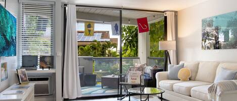 A modern living room with a white sofa, a glass coffee table, and a ceiling fan. There is a sliding glass door leading to a balcony with outdoor furniture.