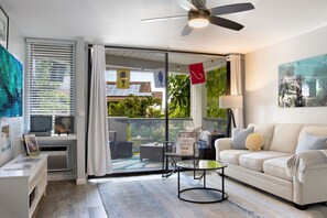 A modern living room with a white sofa, a glass coffee table, and a ceiling fan. There is a sliding glass door leading to a balcony with outdoor furniture. - Modern Kona Retreat • Walk to Ocean & Town (Kailua-Kona)