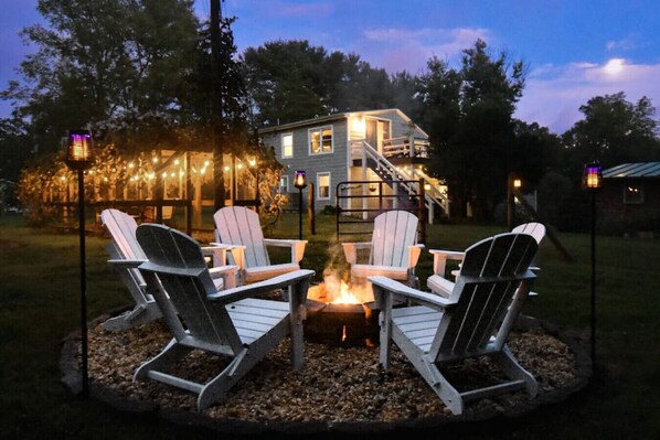 A fire pit with four Adirondack chairs arranged around it, a house in the background, and string lights in the foreground.