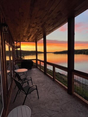 Une terrasse couverte offrant une vue sur un coucher de soleil au-dessus d’un lac, avec des sièges d’extérieur et une rambarde.