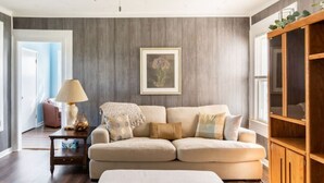 Een woonkamer met een beige bank, een houten kast, een lijst met een foto aan de muur en een lamp op een bijzettafeltje.