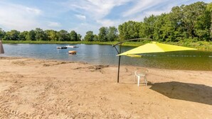 Een zanderig strand met een gele parasol en een witte stoel, een rustig meer en bomen op de achtergrond.