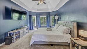 A bedroom with a large bed, a ceiling fan, a TV mounted on the wall, and a window with curtains.