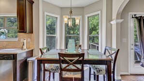 A dining area with a glass table and chairs, a chandelier, and large windows with a view of greenery.