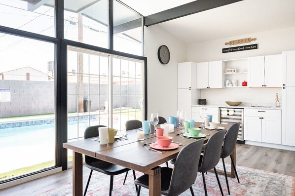 A dining area with a large table set for four, a kitchen in the background, and a view of a swimming pool through large windows.