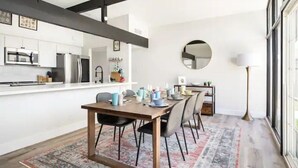 Cozinha moderna com armários brancos, uma mesa de jantar de madeira e cadeiras. Há uma janela grande no lado direito.