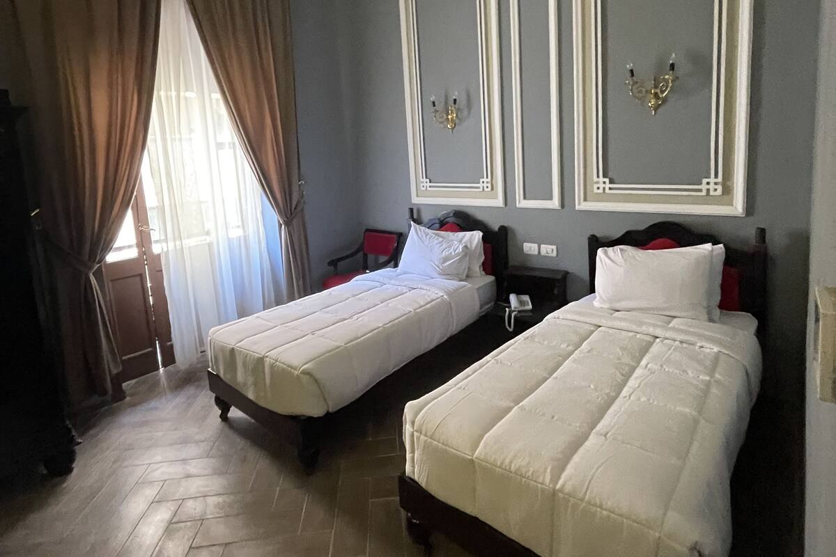 A hotel room with two single beds, a wooden floor, a large window with curtains, and a wall-mounted mirror.