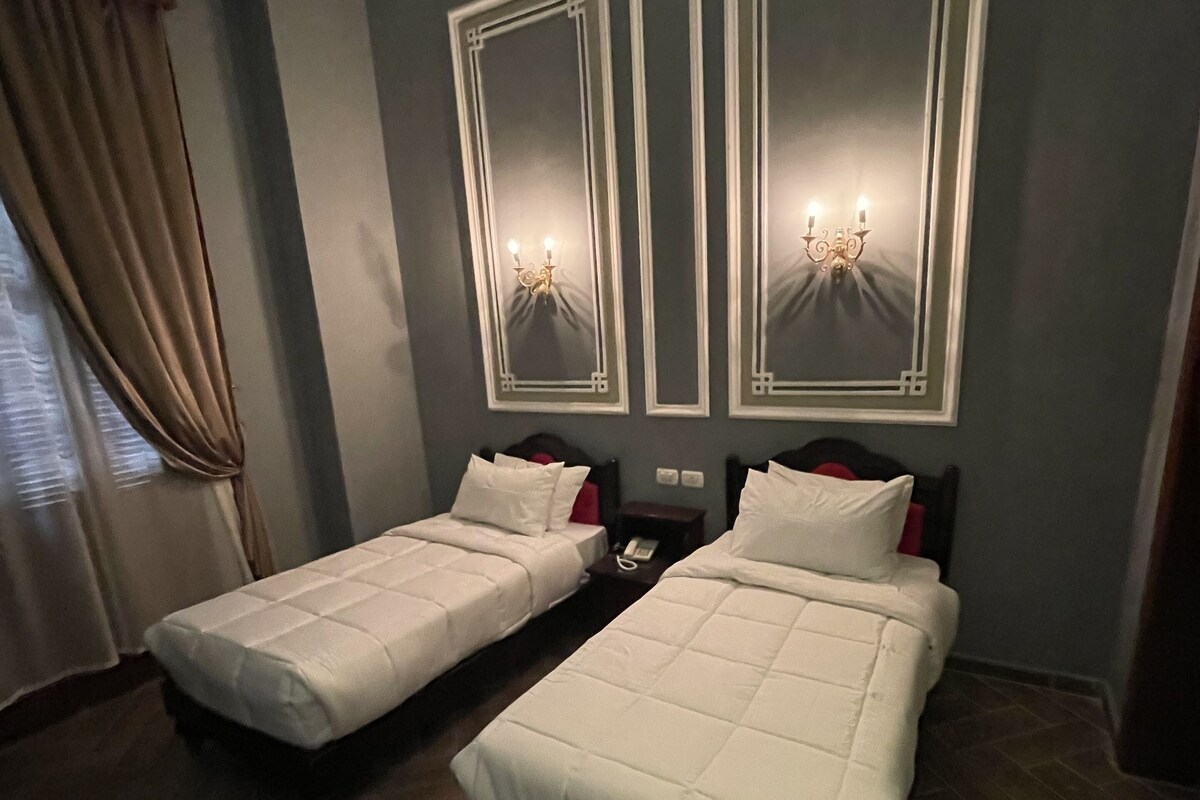 Quarto de hotel com duas camas, uma mesinha de cabeceira, um telefone e uma janela com cortinas.