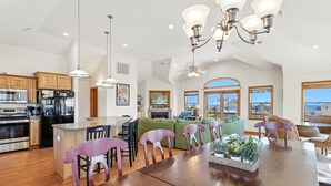 A spacious kitchen with a central island, wooden cabinets, and a large dining table with chairs. - Happy Days: 2-Min Walk Beach, Pool, Hot Tub, 7BR (Southern Shores)