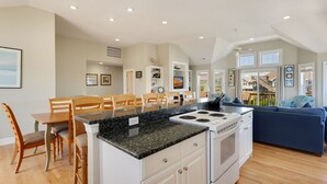 Una cocina amplia con una isla central, muebles blancos y una estufa empotrada. Hay una zona de comedor con sillas de madera y una mesa, y una sala de estar con un sofá azul y vistas al exterior.