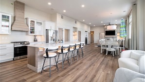 Una cocina moderna con una isla central, taburetes de bar, un comedor con mesa y sillas, y un televisor montado en la pared.