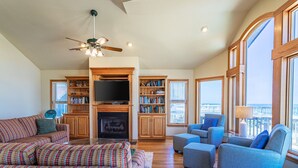 A spacious living room with a fireplace, a ceiling fan, and large windows offering a view. - 6067 The Sand Pebble *Pool *Game Rm *Dog Friendly (Nags Head)