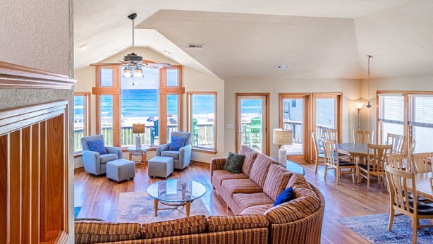 A spacious living room with a large sectional sofa, a glass coffee table, and a dining area with wooden chairs. Large windows offer a view of the beach. - 6067 The Sand Pebble *Pool *Game Rm *Dog Friendly (Nags Head)