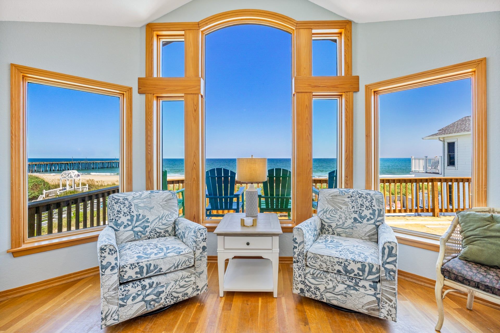 Une chambre avec vue sur la plage, comprenant deux fauteuils à motifs, une petite table blanche et une vue sur l’océan grâce à de grandes fenêtres.
