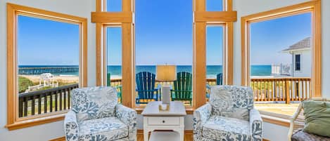 A room with a view of the beach, featuring two patterned armchairs, a small white table, and a view of the ocean through large windows.