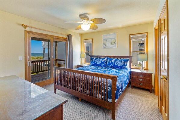 A bedroom with a bed, a desk, a ceiling fan, and a view of the ocean through a door.