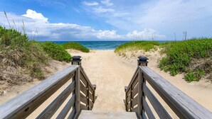 Ein hölzerner Steg führt zu einem Sandstrand, flankiert von grüner Vegetation.