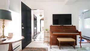 A room with a piano, a television, a round table with a lamp, and a patterned rug. - Locktender Cottage · Historic cottage near Mohonk & Minnewaska, 2BR/2BA (High Falls)
