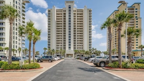 A multi-story hotel with a central entrance, surrounded by palm trees and parked cars. - Neaux Worries: Newly remodeled Ocean Front 16th Floor Pearl Condo! (Navarre Beach)