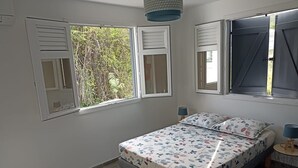 A bedroom with a bed, two windows with shutters, a bedside table with a lamp, and a view of greenery outside. - Magnifique Villa Neuve Avec Piscine (Le Marin)