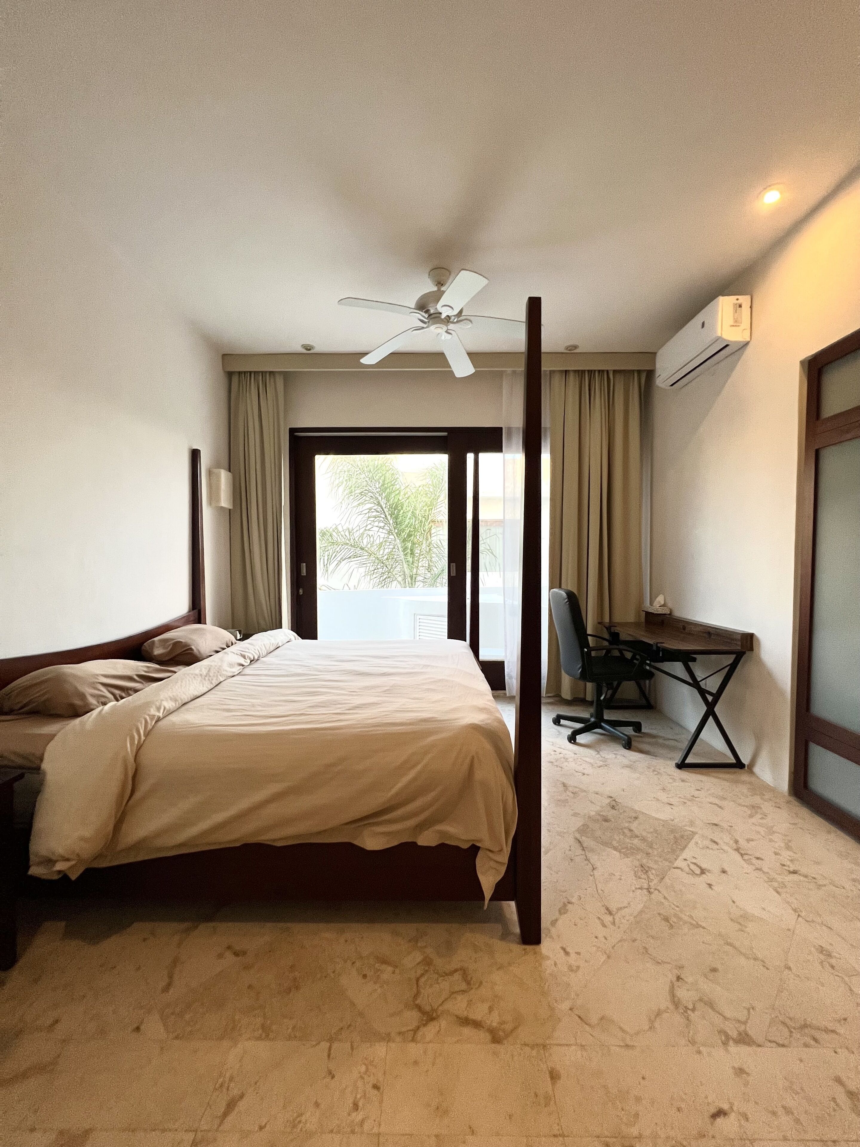 A hotel room with a four-poster bed, a desk with a chair, and an air conditioning unit on the wall.