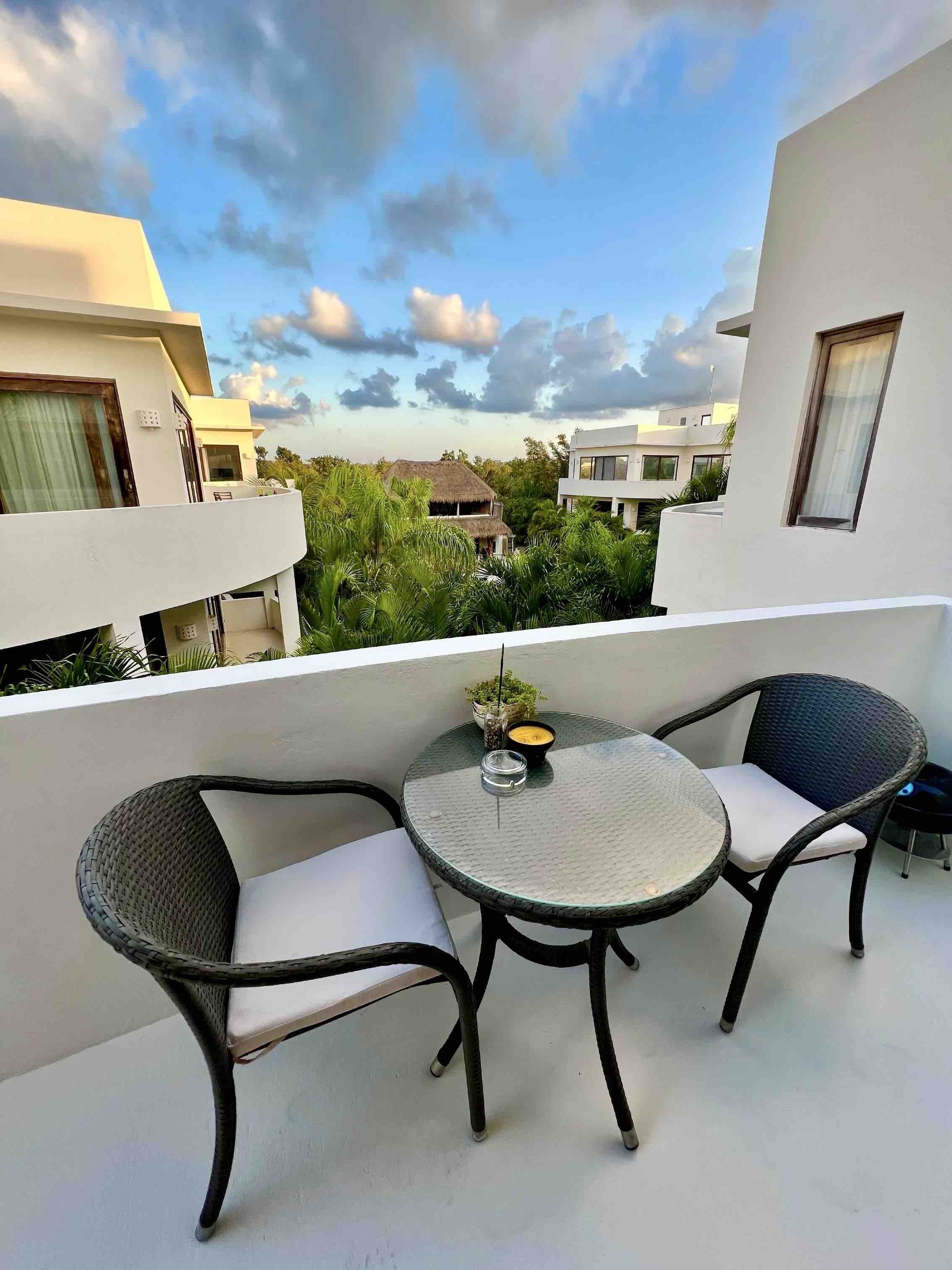 A balcony with two wicker chairs and a small round table, overlooking a residential area with modern houses and lush greenery.