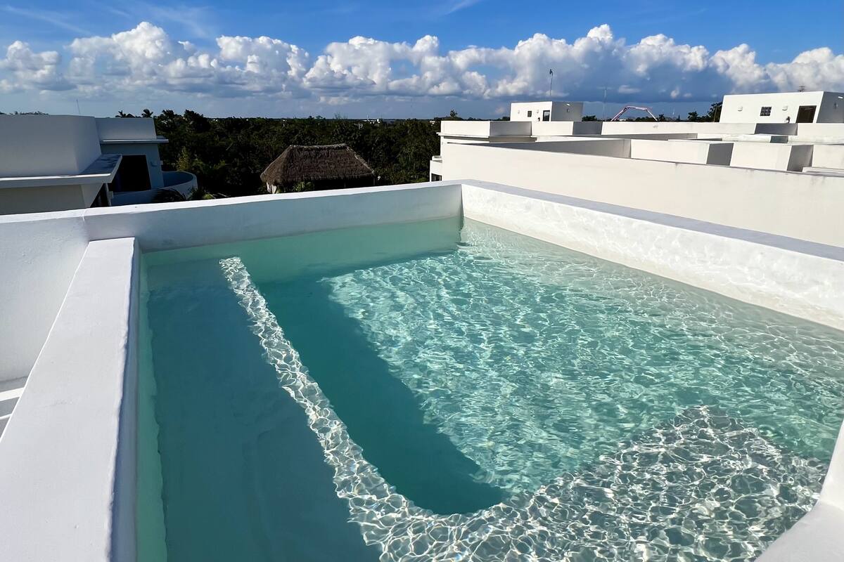 A rooftop swimming pool with clear blue water, surrounded by white buildings and a clear sky.