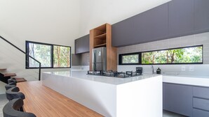 A modern kitchen with a large island, grey cabinets, and a view of trees through the windows. - Carpediem - Casa Luxuosa em Pipa (Tibau do Sul)
