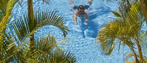 Une piscine entourée de palmiers, avec une personne qui nage et une autre assise au bord de la piscine.