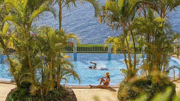 A pool area with palm trees, a stone wall, and a view of the ocean.