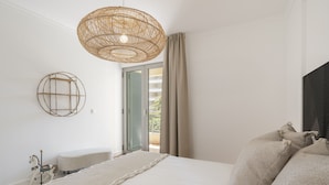 A bedroom with a large bed, a wicker ceiling light, a round wall decoration, a small white chair, and a view of the outside through a window with green shutters.
