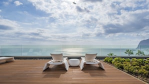 Une terrasse en bois avec deux chaises longues blanches et un pouf rond, d’où l’on peut admirer une mer d’un bleu limpide et une montagne lointaine.