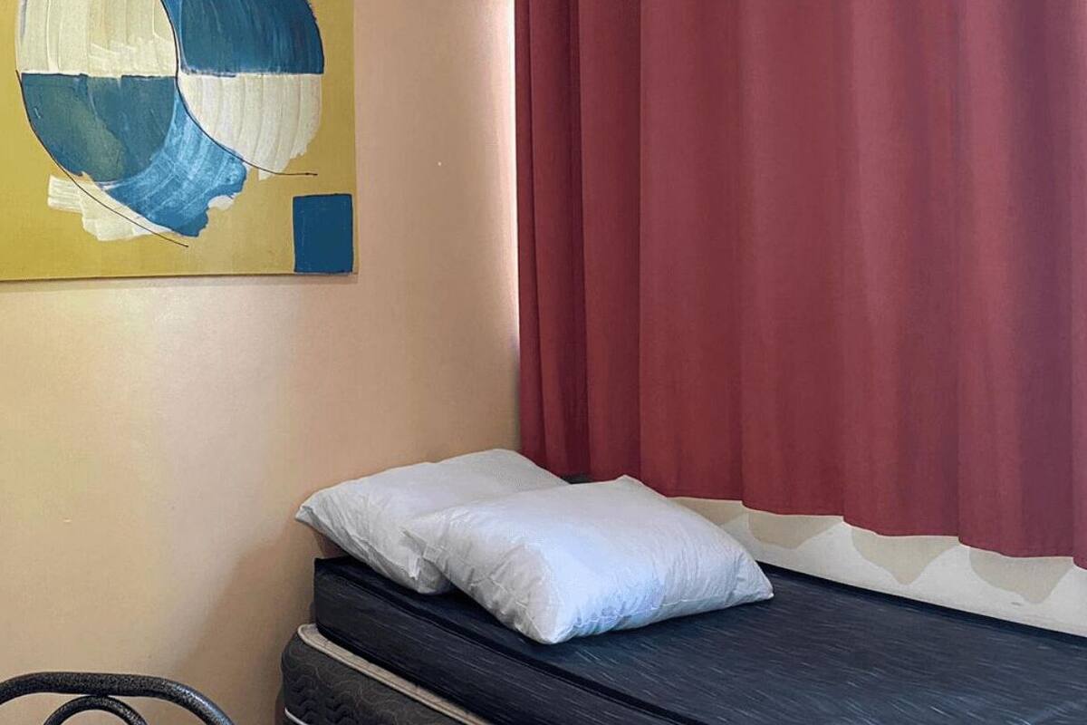 A small, single-bed room with a dark mattress, a white pillow, a wooden shelf, and a painting on the wall.
