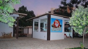 A small, single-story building with a tiled patio area, a wooden table and chairs, and a sign advertising 'Creme Mel Sorveteria'. - Aptos - DiRoma Fiori - Aluguel Economico (Caldas Novas)