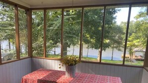 Una habitación con una mesa cubierta por un mantel estampado, una banca y una ventana que da a vistas de árboles y un cuerpo de agua.