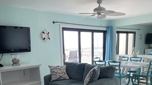 A living room with a grey sofa, a white coffee table, and a dining area with a white table and teal chairs. A ceiling fan is visible, and there is a TV mounted on the wall.