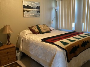 Une chambre à coucher avec un lit, une table de chevet, une lampe, une fenêtre avec des stores et un tableau au mur.