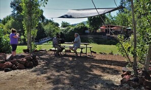 Un espace extérieur aménagé avec des tables de pique-nique et une zone ombragée, entouré d’arbres et offrant une vue sur une maison au loin.